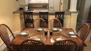 A dining area with a wooden table set for six, surrounded by six chairs. A kitchen with white cabinets and a refrigerator is visible in the background. - Castle Hill Resort Condo (with on-site Aveda Spa) (cavendish)