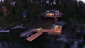 A lakeside cabin with a dock extending into the water, a small boat docked at the end, and a separate cabin with a balcony. - Lake House with Sauna, kayaks, S.U.P.'s, snorkeling sets and boat parking (Polson)