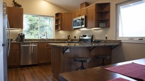 A modern kitchen with wooden cabinets, a stainless steel dishwasher, a microwave, and a granite countertop. There are two bar stools and a window providing natural light. - Lake House with Sauna, kayaks, S.U.P.'s, snorkeling sets and boat parking (Polson)