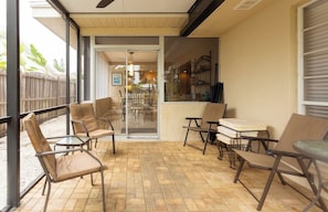 Un patio techado con sillas de mimbre y una mesa, una puerta corrediza de vidrio que da a un espacio interior con una mesa de comedor y sillas, y vistas al exterior.