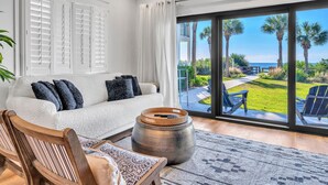 A modern living room with a white sofa, a wooden chair, and a large window offering a view of a well-maintained lawn and palm trees. - Palms A1: Best Prices to Be Ground Level Directly  on the Beach  (Santa Rosa Beach)