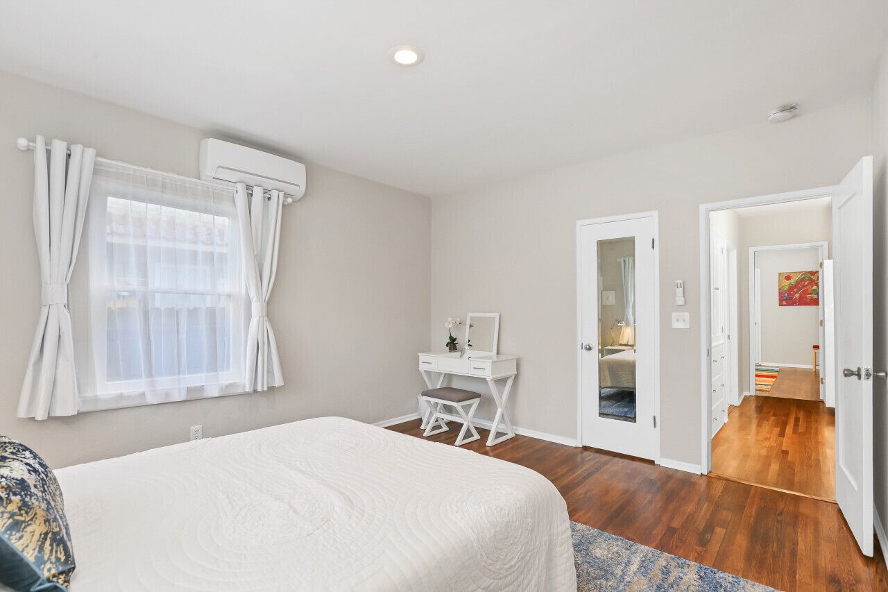 Une chambre à coucher avec un lit, un bureau et une chaise. Il y a une fenêtre avec des rideaux, une porte qui donne sur une autre pièce et un couloir.