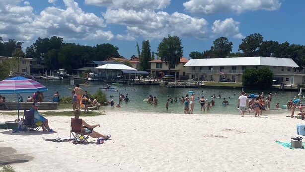 A beach with people swimming and playing, a dock with boats, and buildings in the background. - Super Clean and Super Comfortable with boat ramp under half mile down the road. (Crystal River)