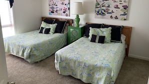 A bedroom with two twin beds, each with matching green and blue patterned bedding, a green nightstand, and two beach-themed wall art pieces.