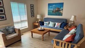 A cozy living room with a wicker sofa, a blue couch, and a wooden coffee table. There are framed pictures on the wall and a painting of a beach scene.