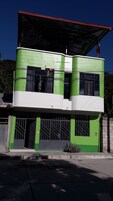Un edificio de dos pisos con fachada verde y blanca, ventanas grandes y un porche cubierto.