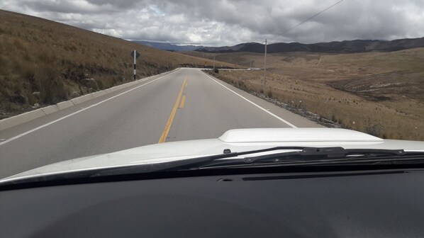 Una carretera con un coche blanco al frente, rodeada de hierba seca y montañas al fondo.