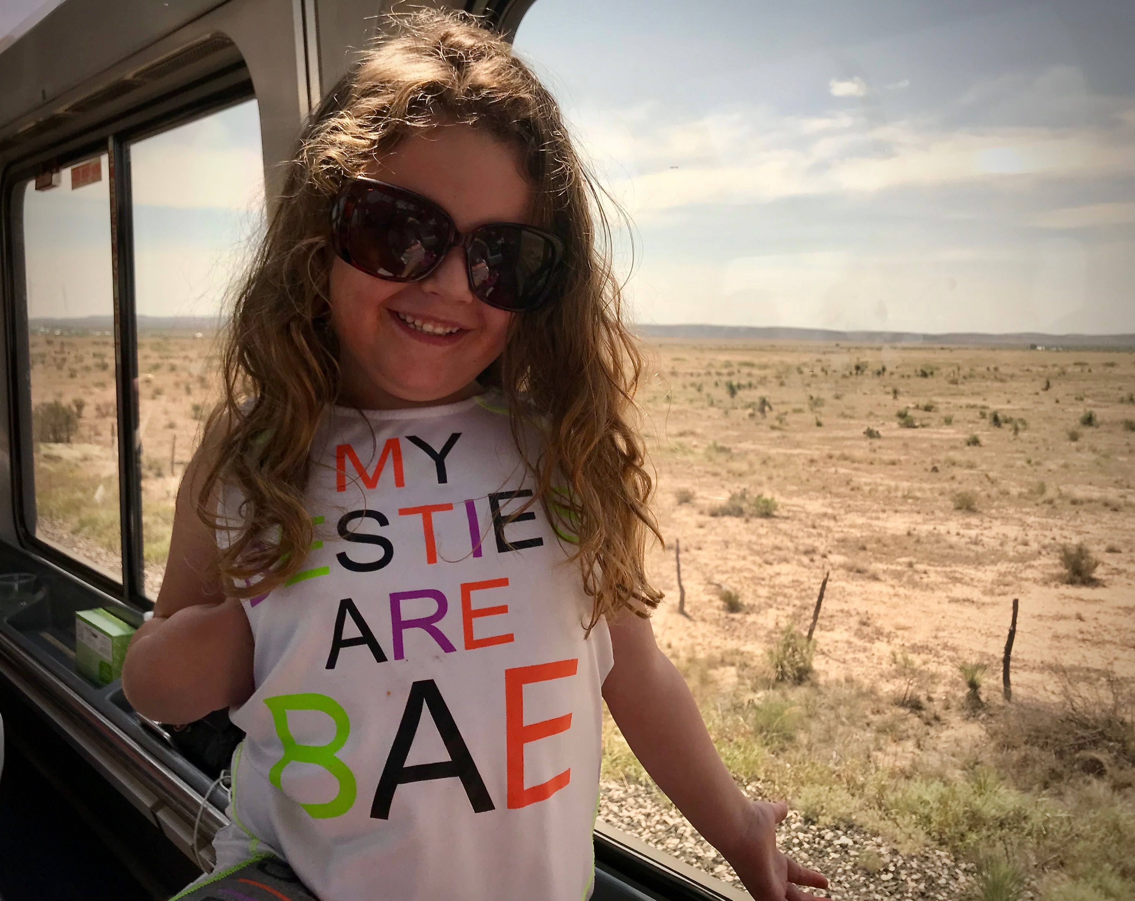 A person wearing a white t-shirt with colorful text, standing by a window with a view of a dry landscape.