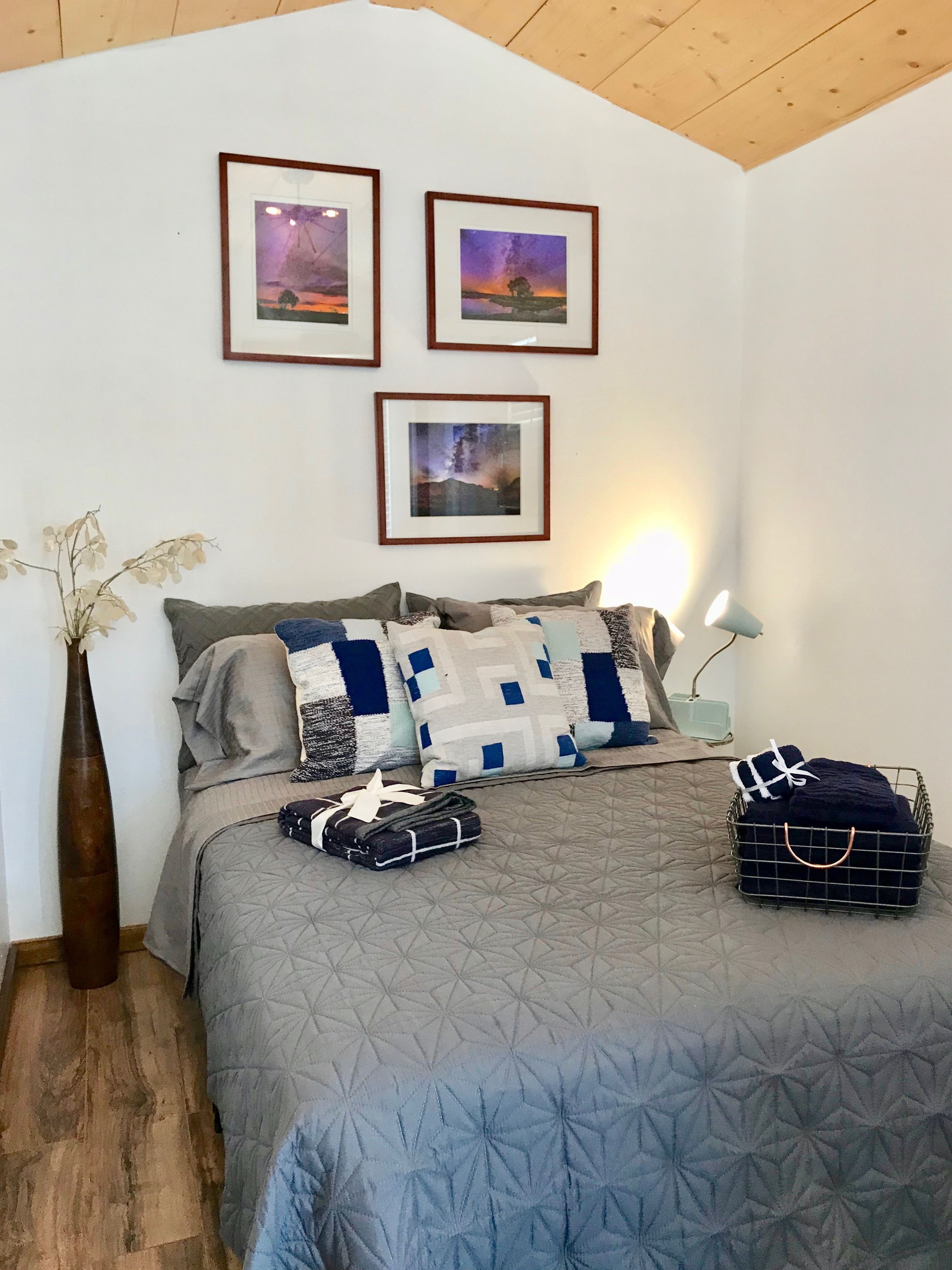 Un dormitorio con una cama, almohadas, un jarrón, una lámpara y tres cuadros en la pared.