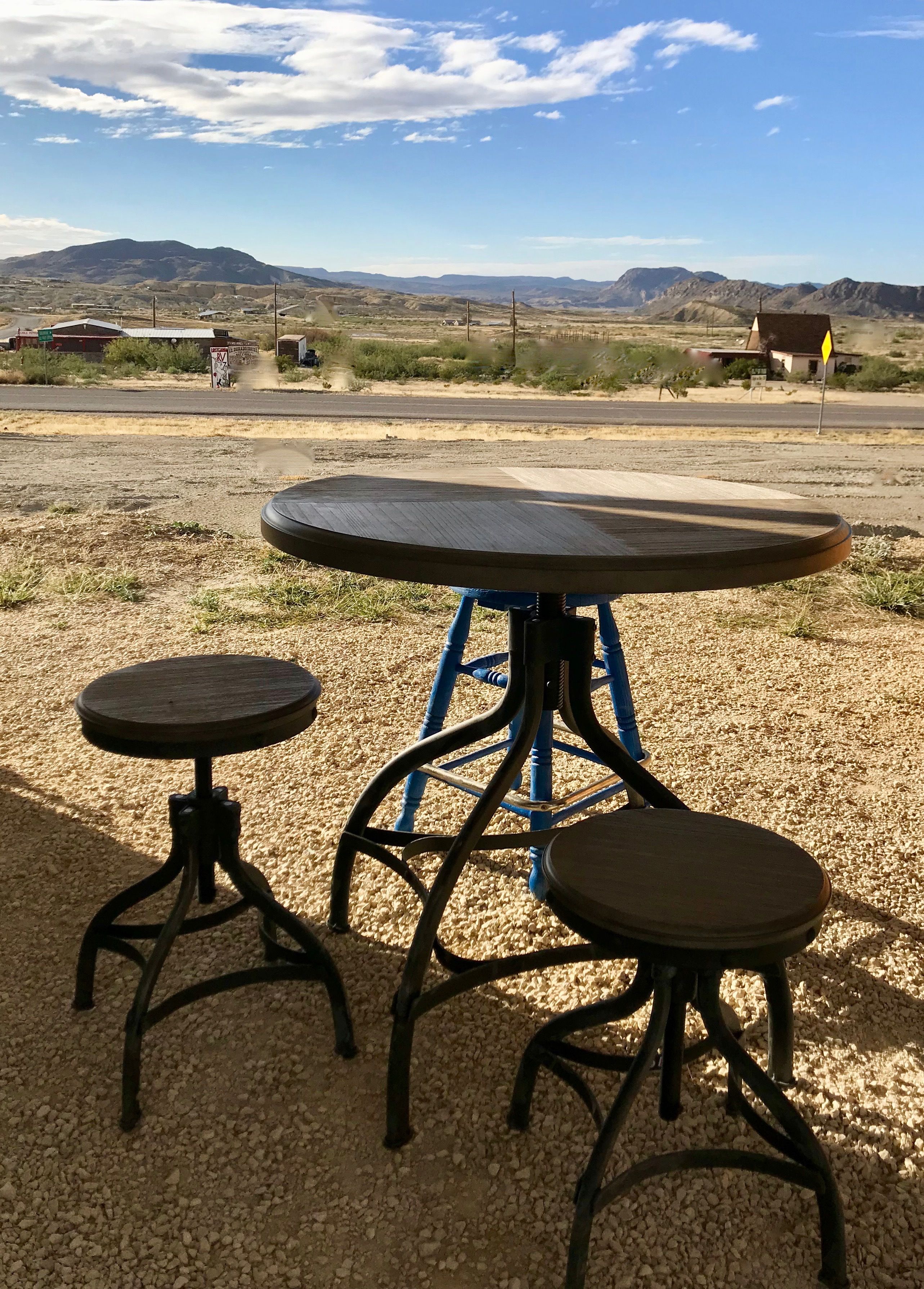 A rustic outdoor seating area with a round table and two stools, set against a backdrop of a clear sky and distant mountains.