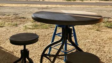 A rustic outdoor seating area with a round table and two stools, set against a backdrop of a clear sky and distant mountains.