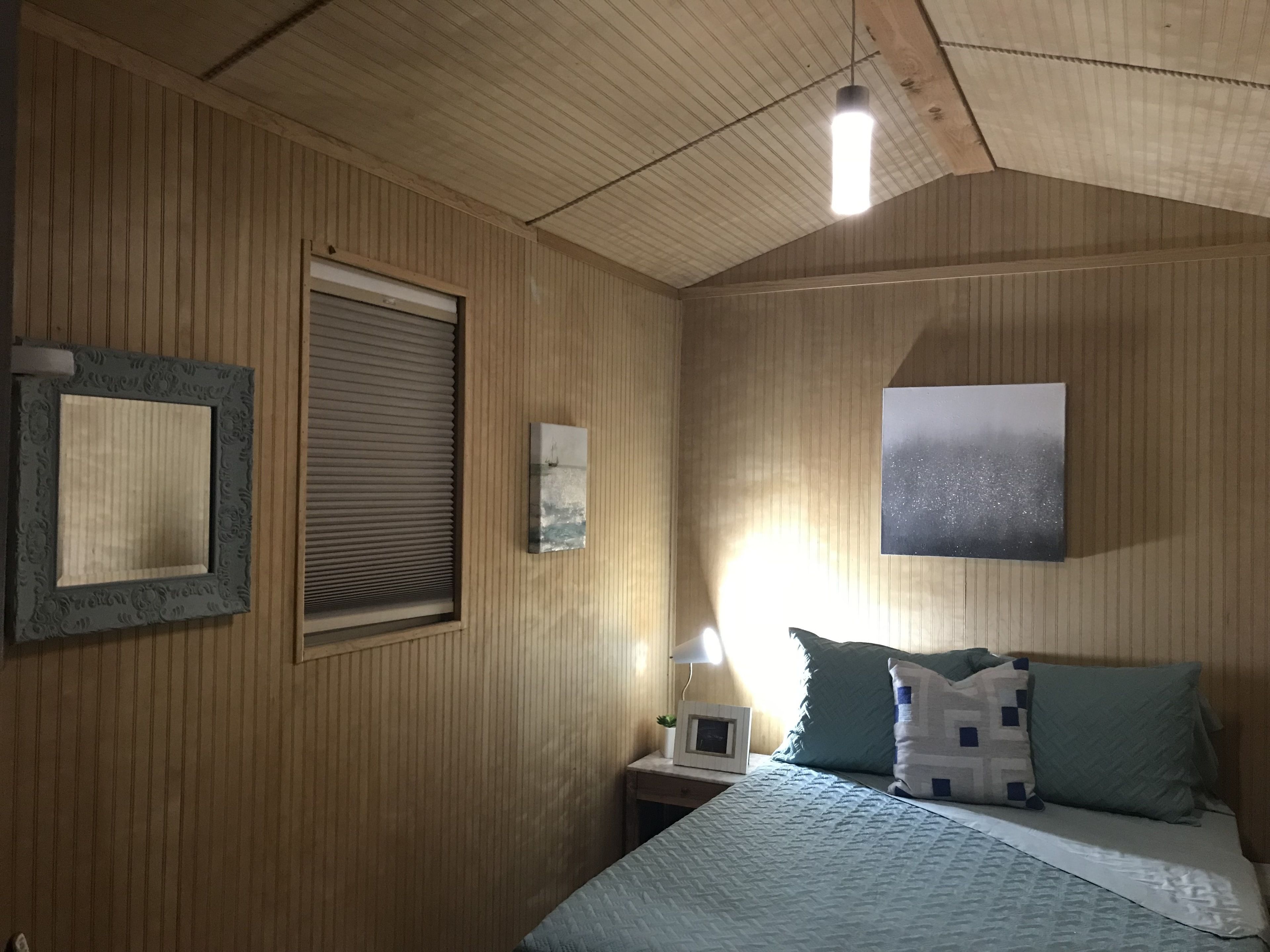A room with wooden paneling, a bed with a quilt, a bedside table with a lamp, a framed picture, and a window with blinds.