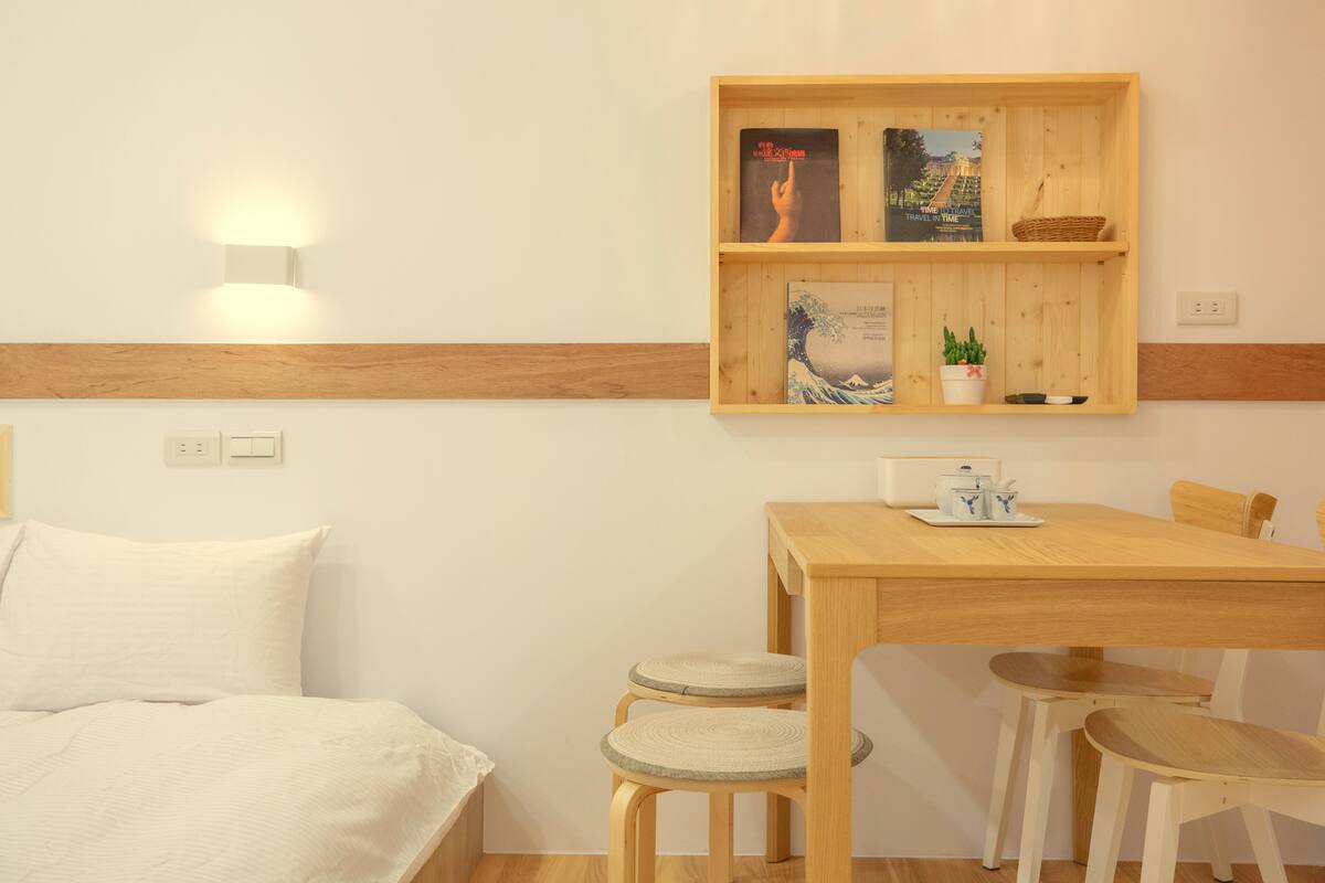 A room with a bed, a wooden table with chairs, a shelf with books and a plant, and a wall-mounted light.
