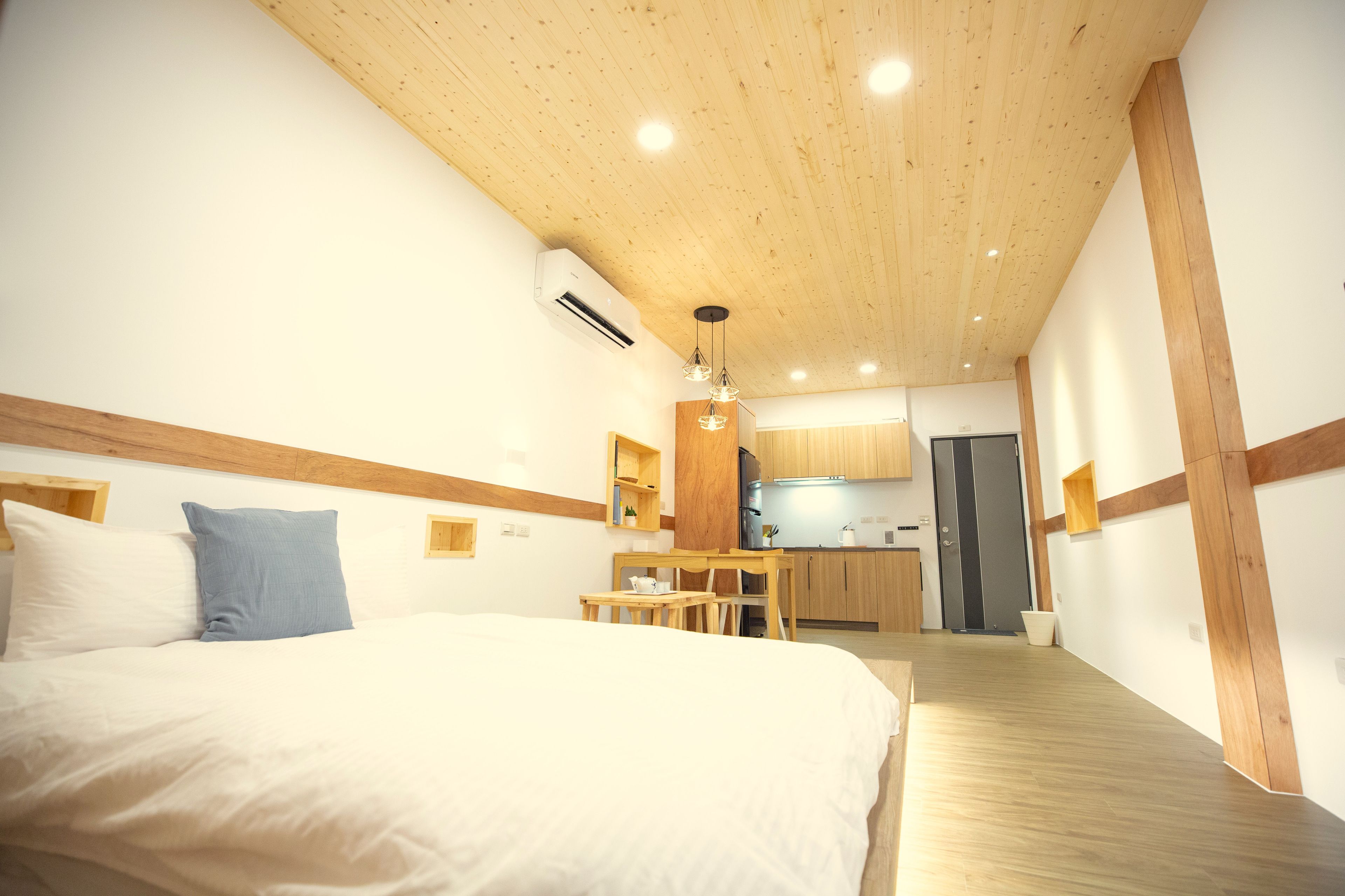 A bedroom with a wooden ceiling, a bed with white and blue pillows, a wooden desk, and a kitchen area in the background.