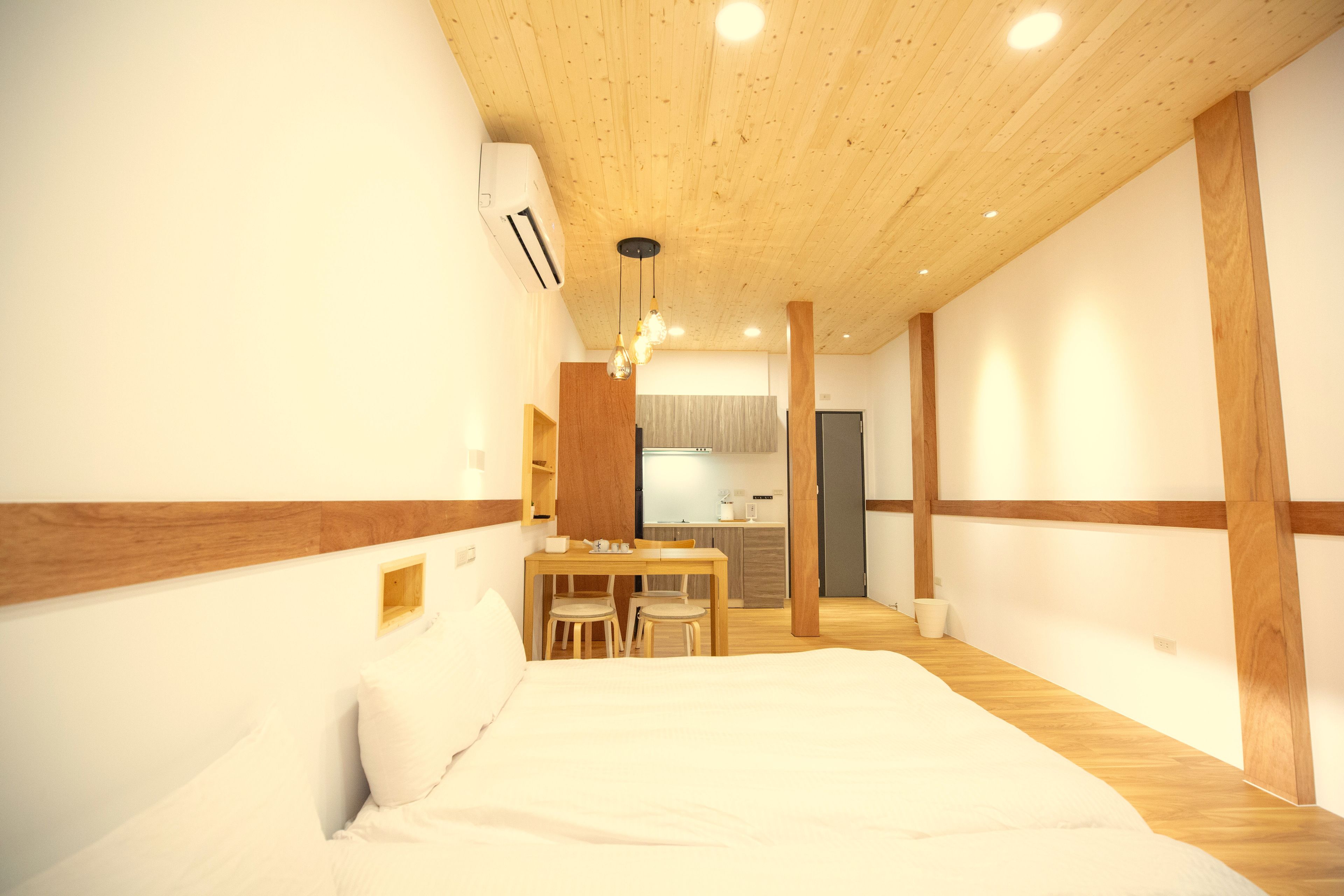 Quarto com teto de madeira, cama, escrivaninha com cadeiras e uma kitchenette ao fundo.