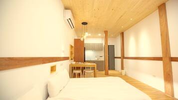 Quarto com teto de madeira, cama, escrivaninha com cadeiras e uma kitchenette ao fundo.