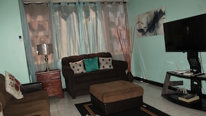 A living room with a brown sofa, teal cushions, a black ottoman, a TV on a stand, and a lamp. - New Kingston Serenity at The Oxford (Kingston)