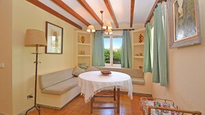 A cozy room with a wooden ceiling, a round table with a white tablecloth, a bench with cushions, a floor lamp, and a window with curtains. - Rustic - Cozy country house with pool in Mancor de la Vall, Mallorca (Inca)