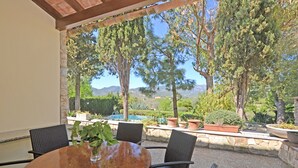 An outdoor patio with a round table and chairs, surrounded by potted plants and a view of a swimming pool and mountains. - Rustic - Cozy country house with pool in Mancor de la Vall, Mallorca (Inca)