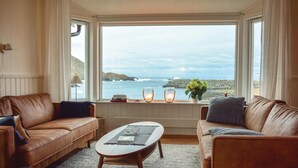 Ein gemütliches Wohnzimmer mit zwei Ledersofas, einem Couchtisch und einem großen Fenster mit Blick auf das Meer und die felsige Küste.