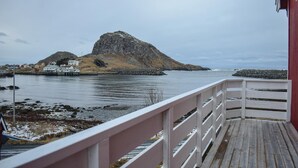 Blick auf die Küste von einer Holzdielen-Terrasse mit weißer Reling, mit Blick auf eine felsige Insel mit einer kleinen Siedlung und einem Steg, der sich in ruhiges Wasser erstreckt.