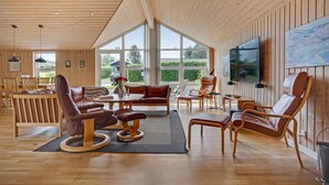 A spacious living room with wooden floors and walls, large windows, a flat-screen TV, and various seating options. - Nice home in Ebeltoft with kitchen (Ebeltoft)