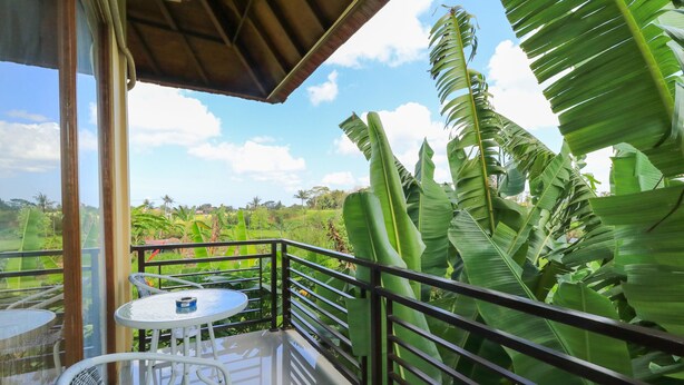 A balcony with a glass door, white wicker furniture, and a view of a lush green landscape. - Sunari Guest House (Canggu)