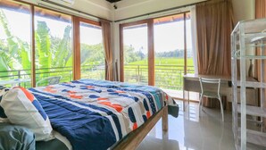 A bedroom with a bed, a desk, and a chair. There are large windows with curtains, and a view of greenery outside. - Sunari Guest House (Canggu)