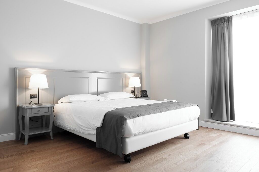 A modern hotel room with a large bed, two bedside tables with lamps, a nightstand, and a window with curtains.