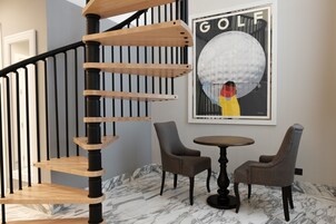 Moderni portaat, jossa on puiset askelmat ja mustat kaiteet, kehystetty golf-aiheinen taideteos sekä kaksi verhoiltua tuolia pienen pyöreän pöydän ympärillä marmorilattialla.