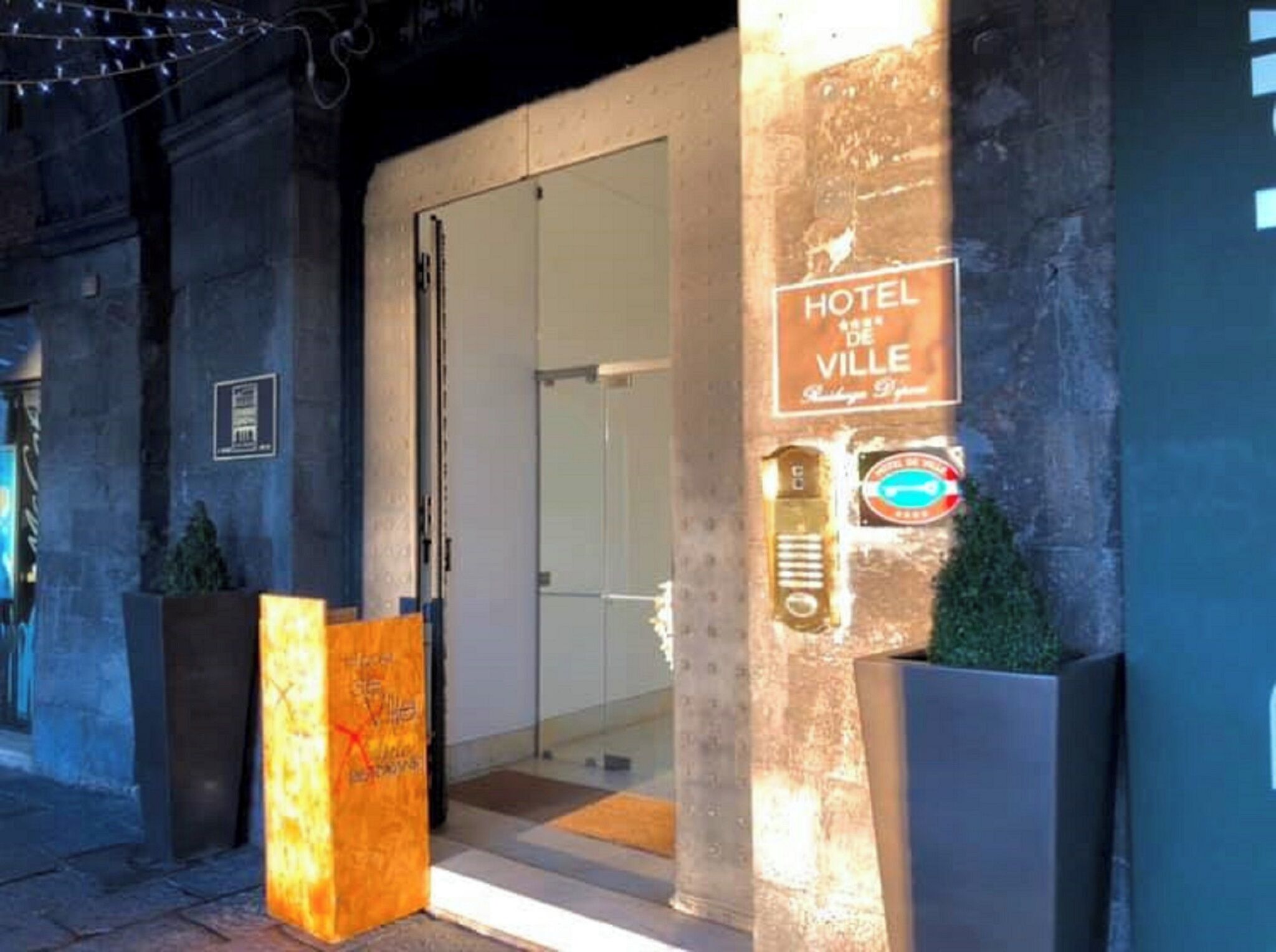 The entrance to Hotel de Ville with a glass door, a signboard, and a large planter.