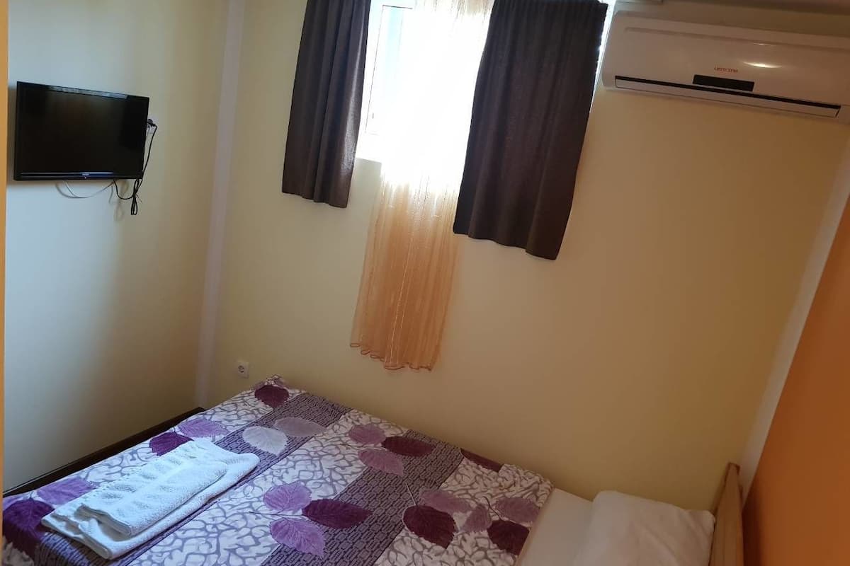 A small, single-bed room with a bed, a TV mounted on the wall, and an air conditioner.