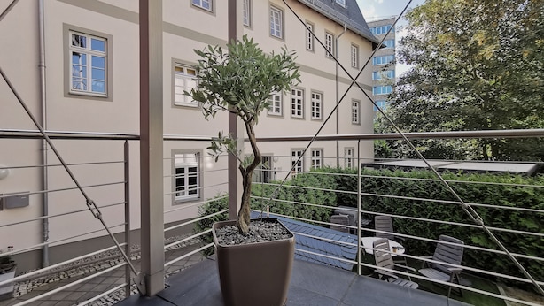En balkon med en potteplante, gelænder i metal og udsigt til en bygning med mange vinduer.