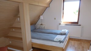 Et soveværelse med køjeseng i træ, et lille natbord med lampe, en radiator og et vindue med udsigt til grønne omgivelser.