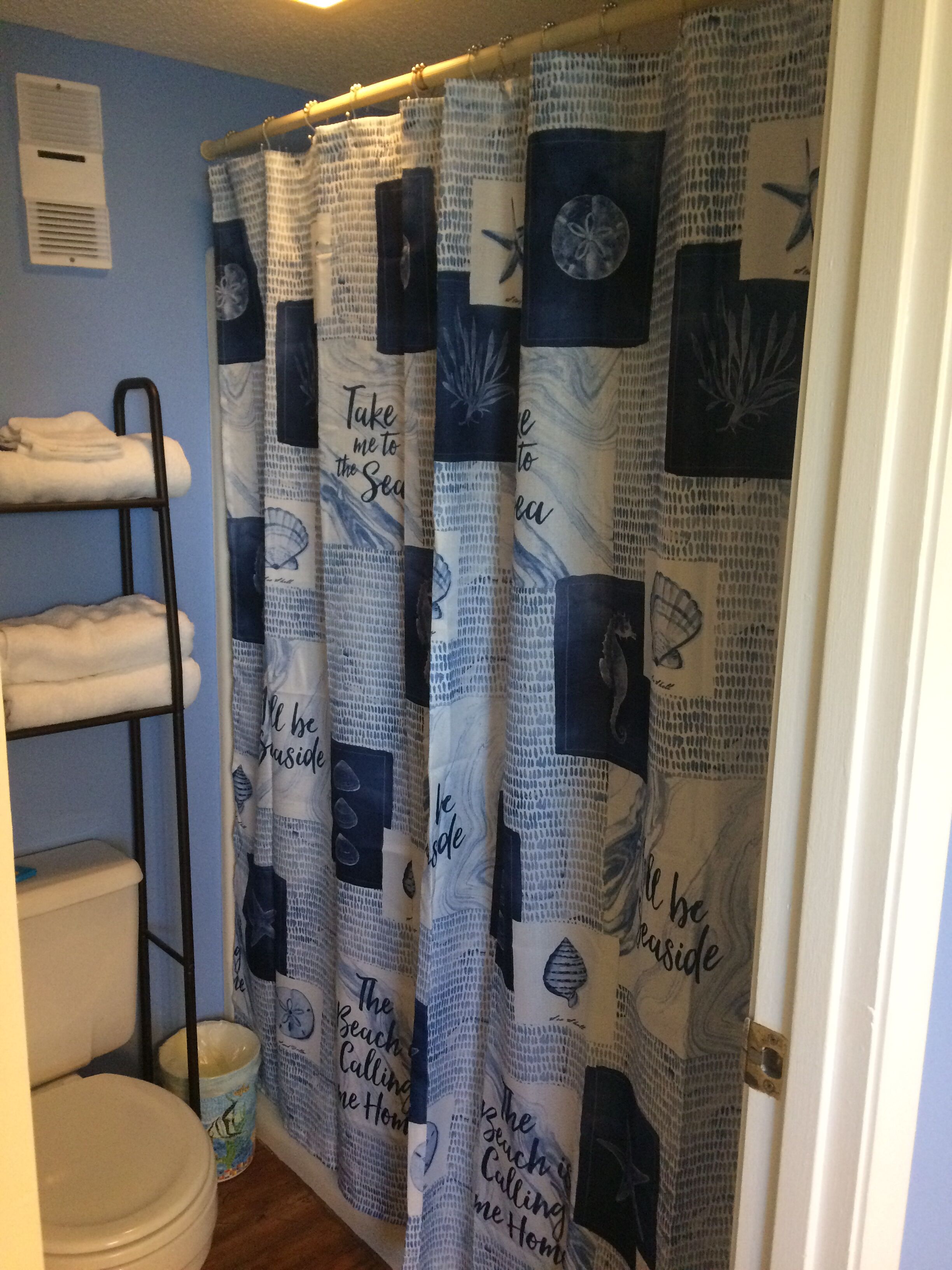 A bathroom with a shower curtain featuring nautical designs and text, a towel rack with folded towels, a toilet, and a trash can.