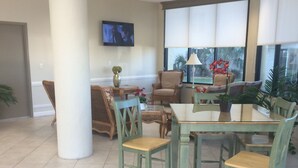 Una habitación amplia con un gran ventanal, un televisor de pantalla plana en la pared y una zona de comedor con sillas verdes y una mesa.