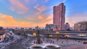 A cityscape with a tall building, a roundabout, and two cups of coffee on a tray. - Tahrir Plaza Suites (Cairo)