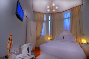 A hotel room with a bed, bedside tables, a TV mounted on the wall, and a chandelier.