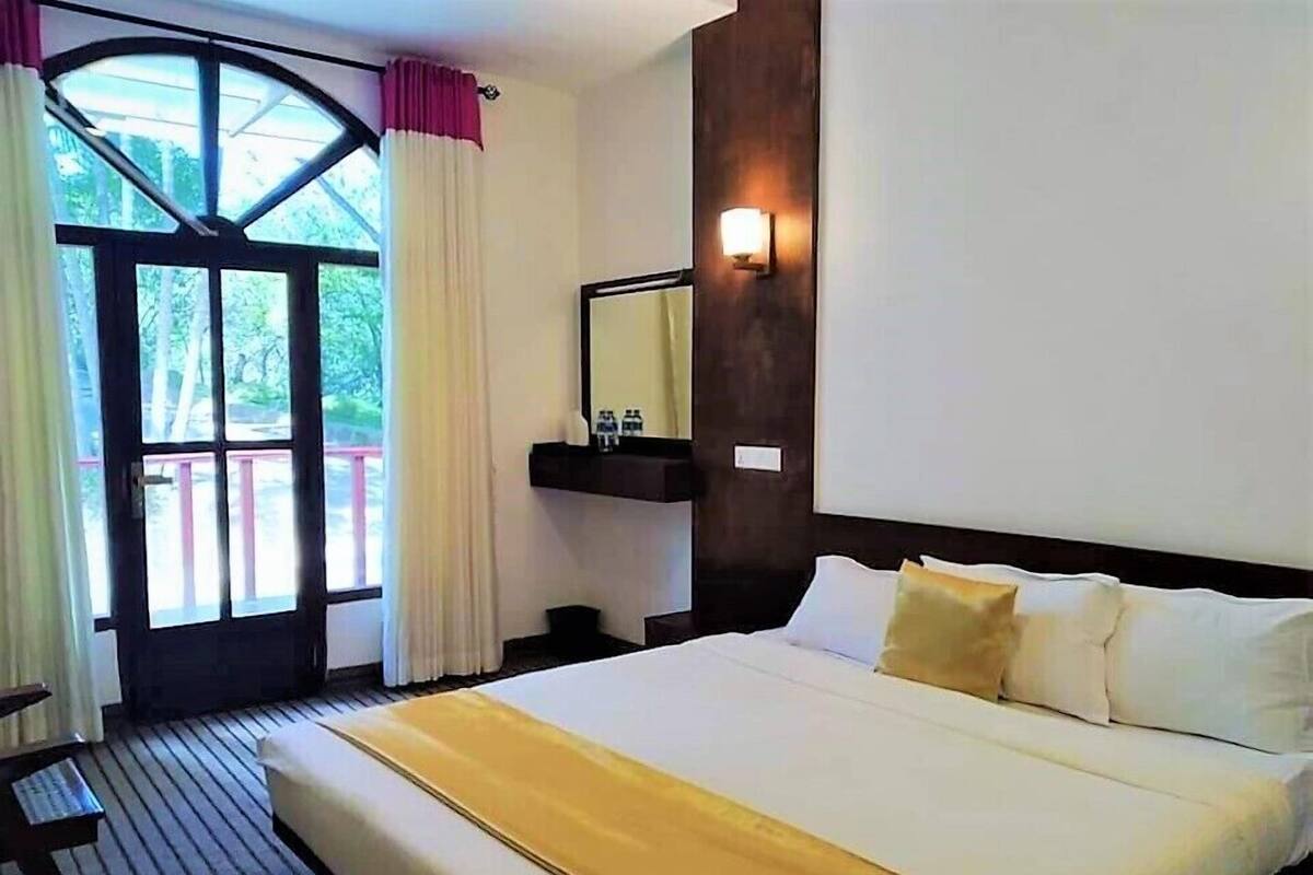 A hotel room with a large bed, a wooden floor, a window with curtains, a mirror, and a nightstand with a lamp.