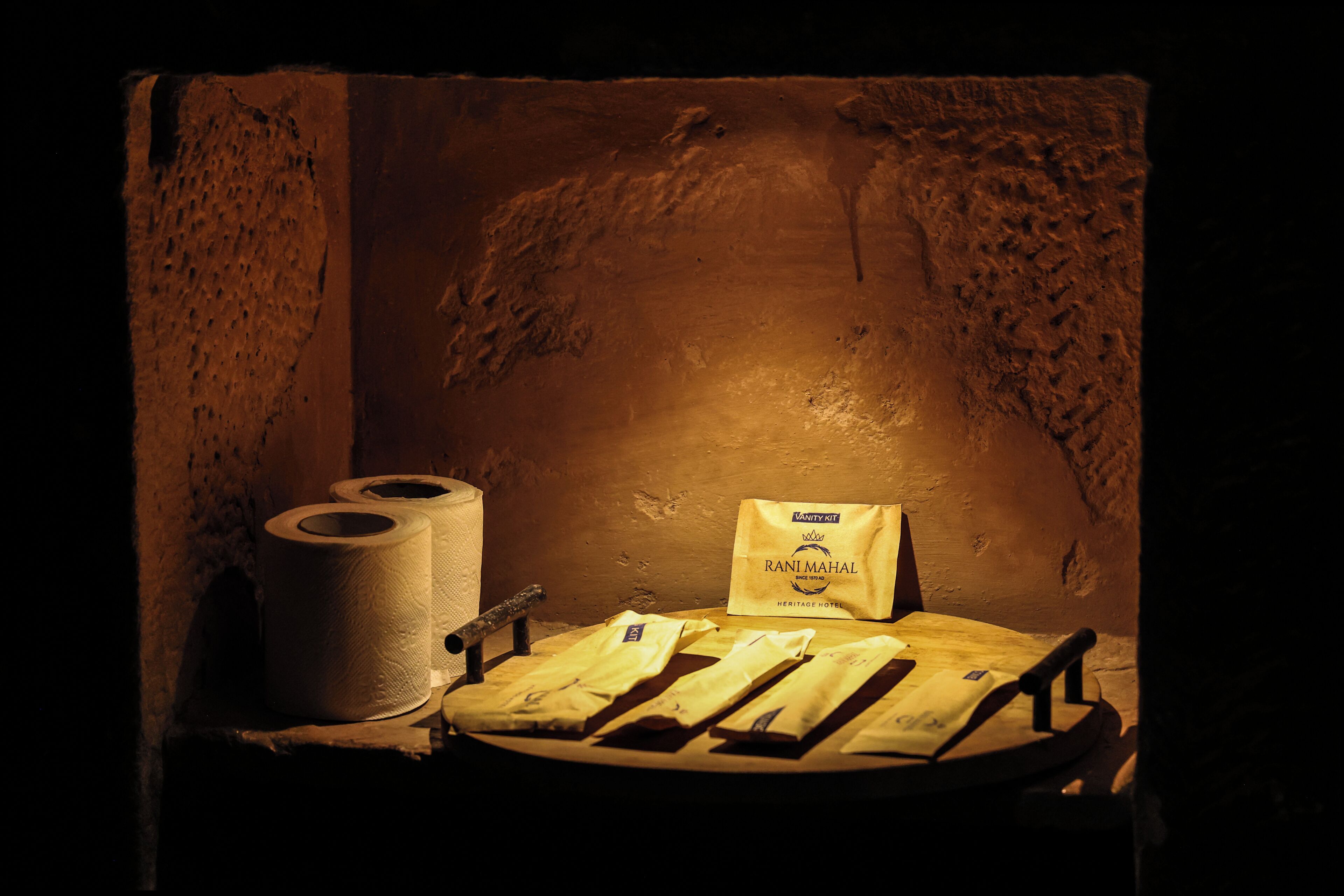 A dimly lit room with a textured wall, a roll of toilet paper, and a bag labeled 'Rani Mahal Heritage Hotel.'