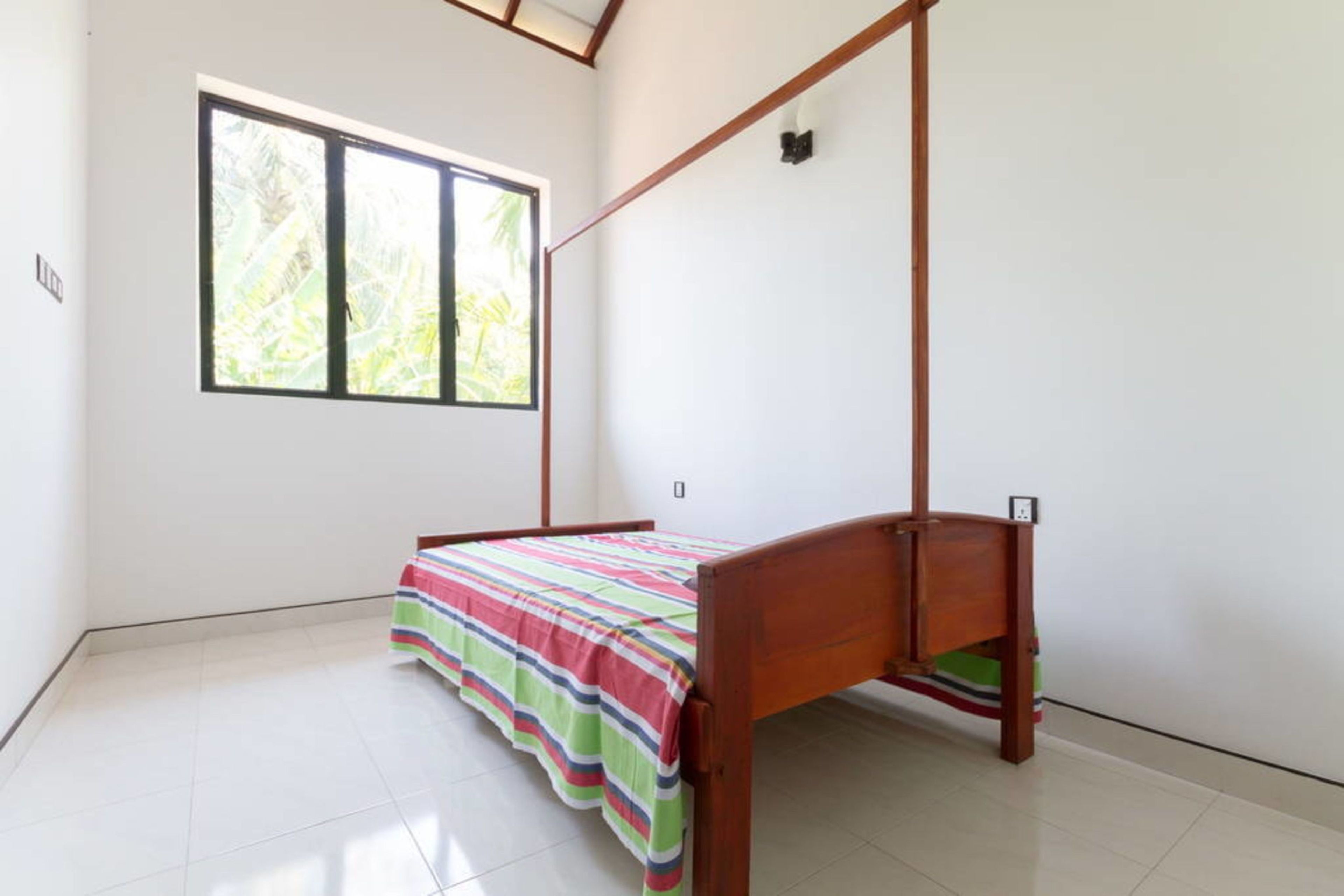 A single bed with a wooden frame and a colorful striped bedspread in a room with white walls and a large window.