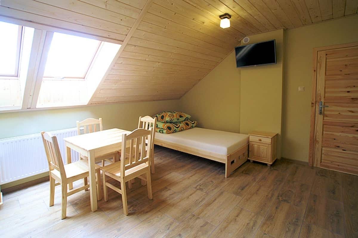 Una habitación pequeña con paneles de madera, techo inclinado, una cama, un televisor montado en la pared y una mesa de comedor con sillas.