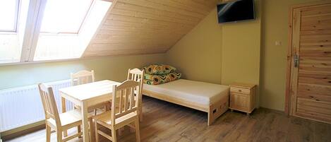 A small, wooden-paneled room with a sloped ceiling, a bed, a TV mounted on the wall, and a dining table with chairs.
