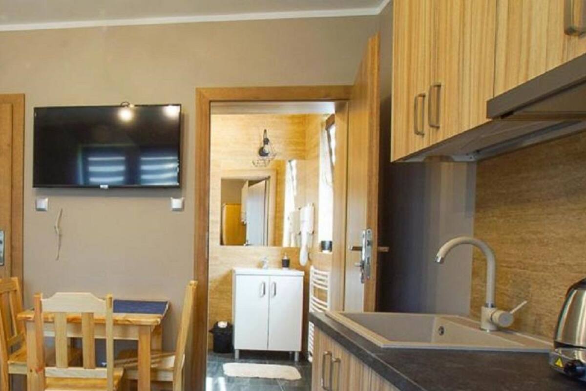 Una cocina compacta con armarios de madera, un televisor integrado, un pequeño comedor con mesa y sillas, y un baño a la vista con lavamanos y espejo.