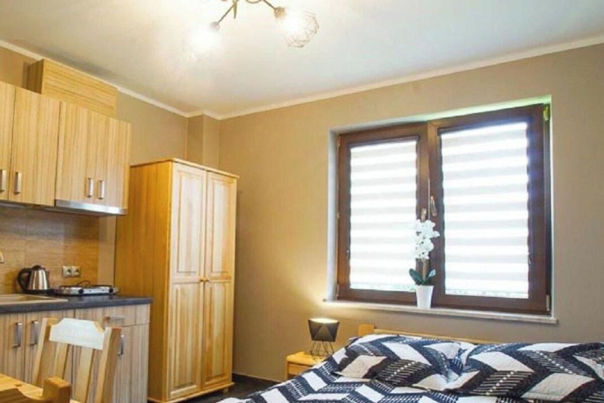Quarto pequeno com cozinha compacta, uma cama com colcha estampada e uma janela com persianas.