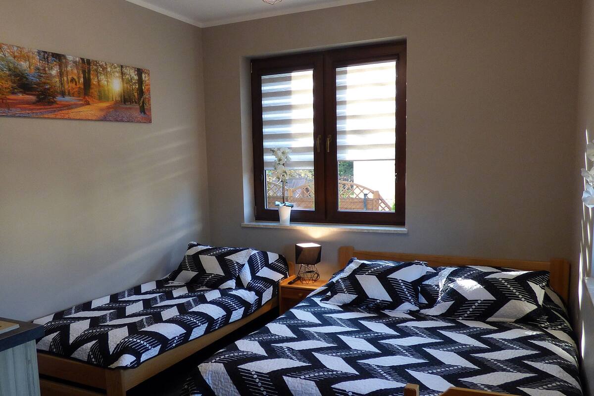 Un dormitorio con dos camas individuales, un cuadro en la pared, una ventana con persianas y una mesita con una lámpara.