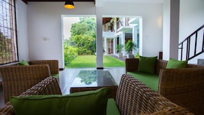 Un vaste espace intérieur aménagé avec des meubles en osier et une table en verre, qui donne sur un jardin luxuriant et un bâtiment à plusieurs niveaux.