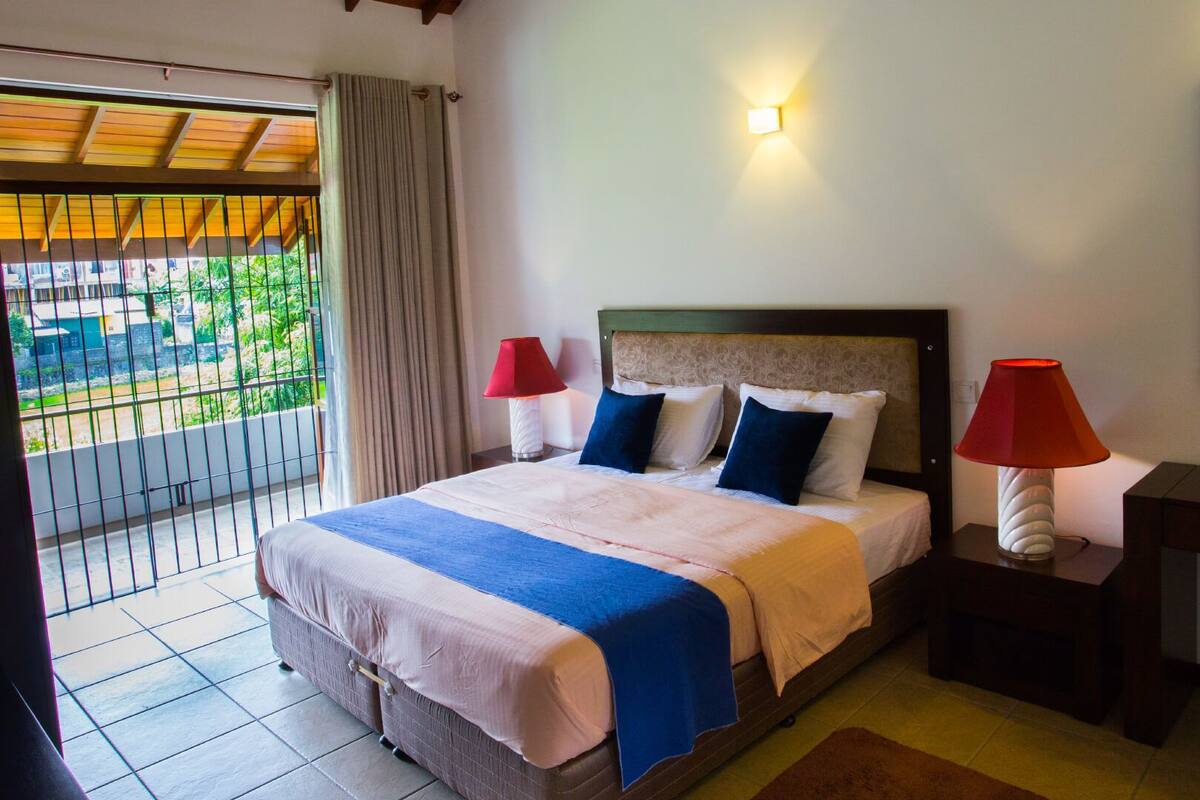 Habitación de hotel con cama, dos lámparas de noche, mesita de noche, espejo y vistas a zonas verdes.