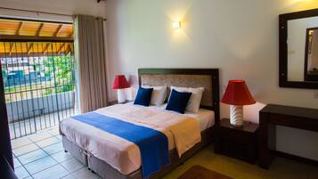 A hotel room with a bed, two bedside lamps, a nightstand, a mirror, and a view of greenery outside.