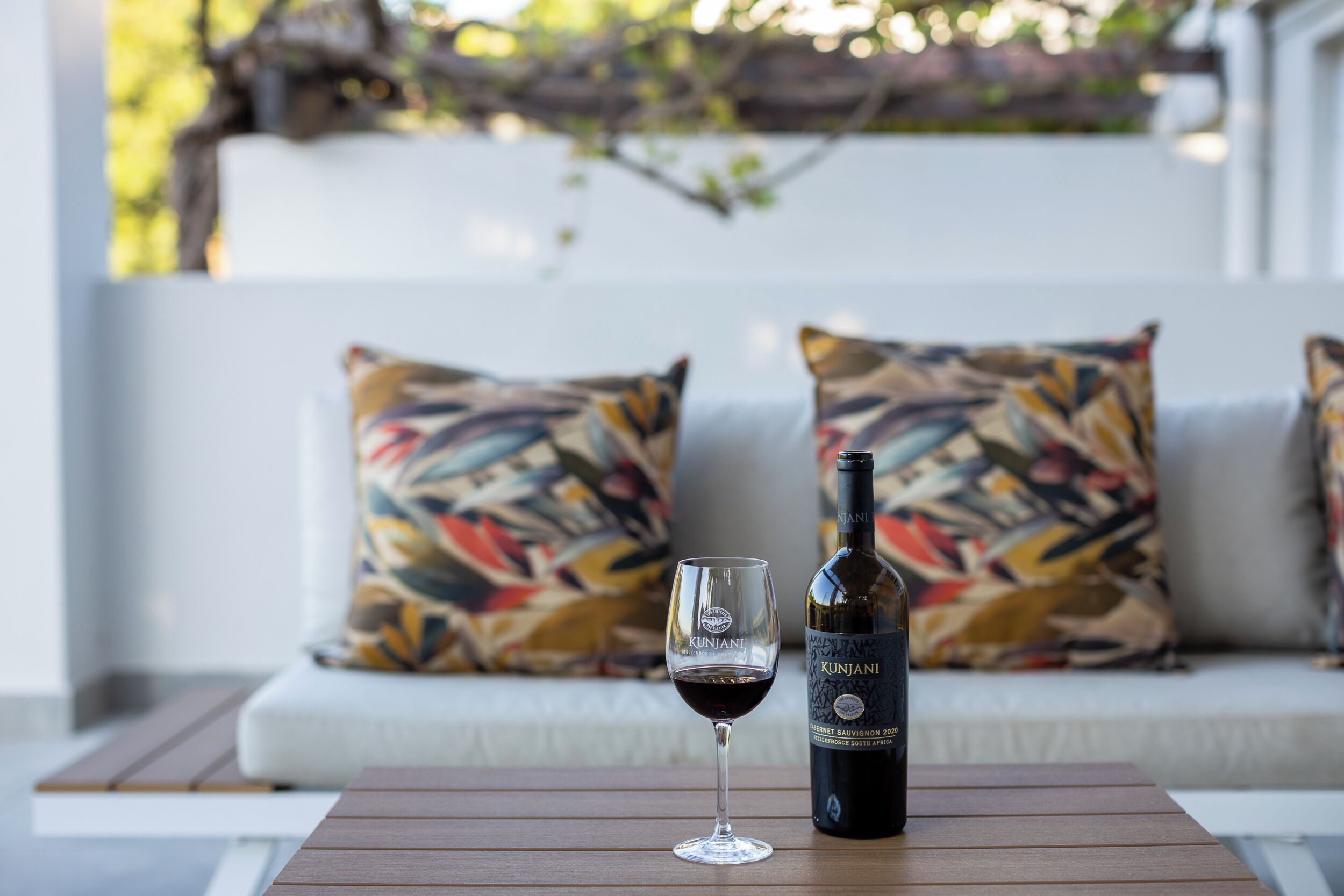 A bottle of Kunjani Cabernet Sauvignon 2020 and a glass of red wine on a wooden table in front of a white sofa with colorful pillows.